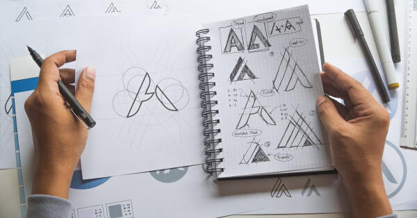 Designer skizziert Logo-Entw&uuml;rfe mit Stift in Notizbuch und auf Papierb&ouml;gen
