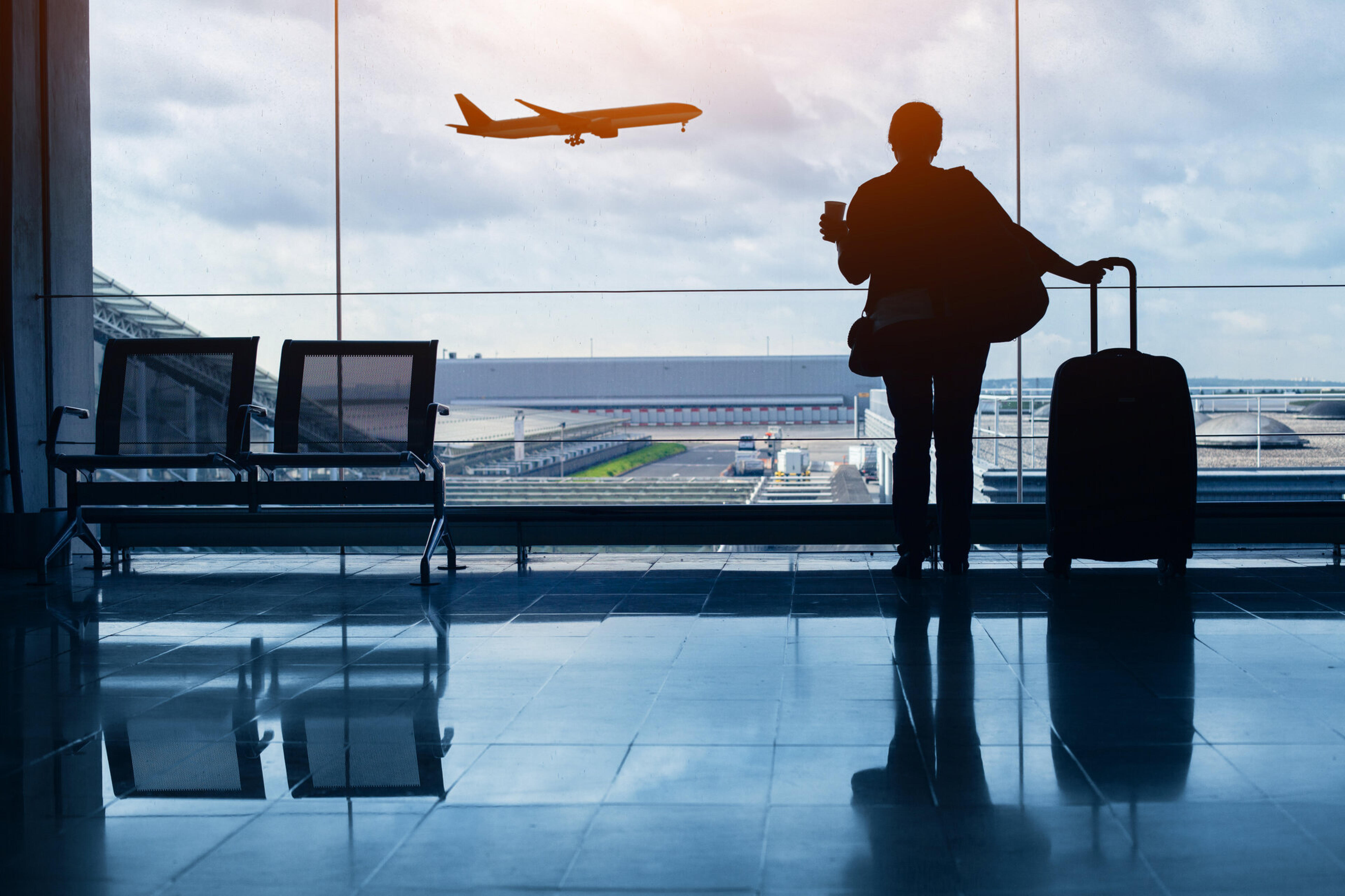 Reisender mit Koffer am Flughafenfenster, im Hintergrund startet ein Flugzeug