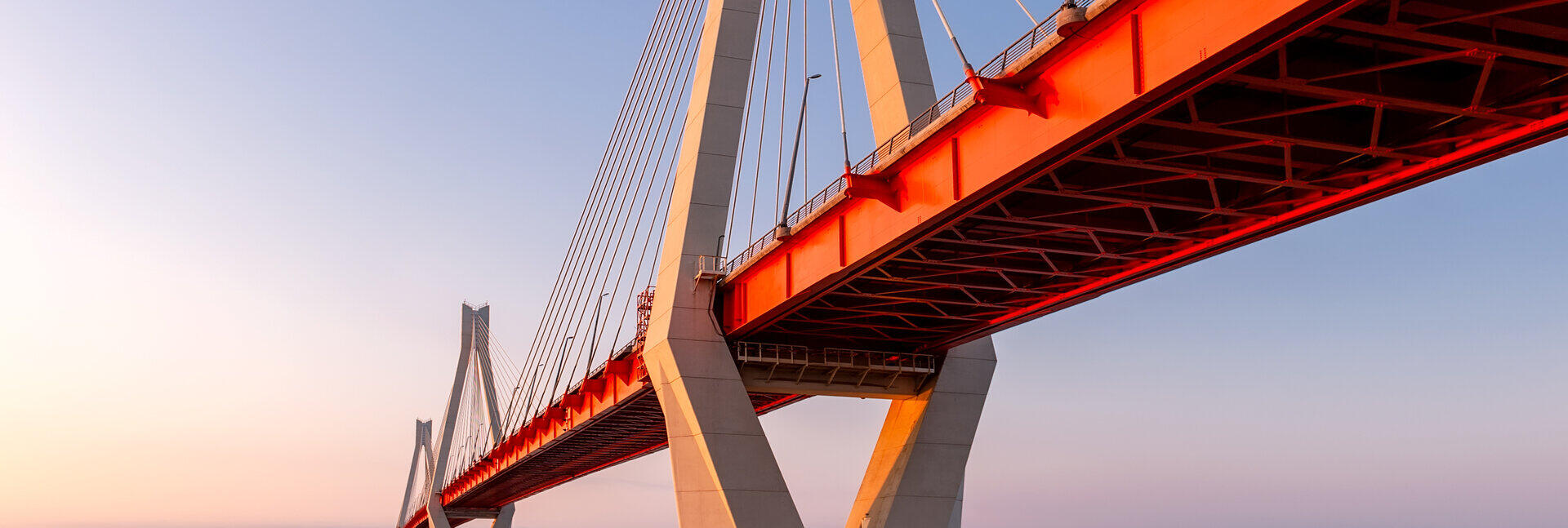 Schr&auml;gseilbr&uuml;cke mit wei&szlig;em Pylon und rotem Fahrbahntr&auml;ger von unten vor klarem Abendhimmel