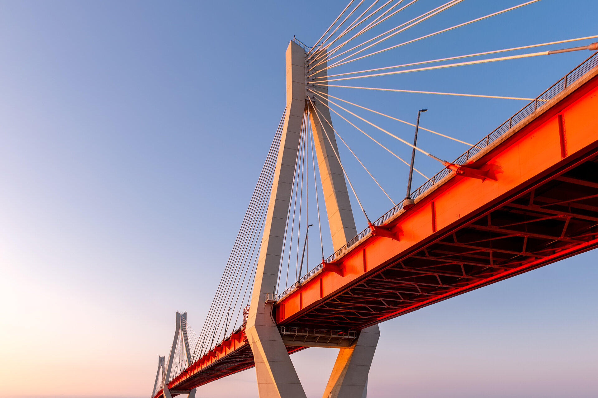 Cable-stayed bridge with white pylon and red deck seen from below against a clear evening sky