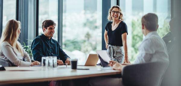 Team im Konferenzraum mit Glasfront, eine stehende Frau spricht lachend mit Kollegen