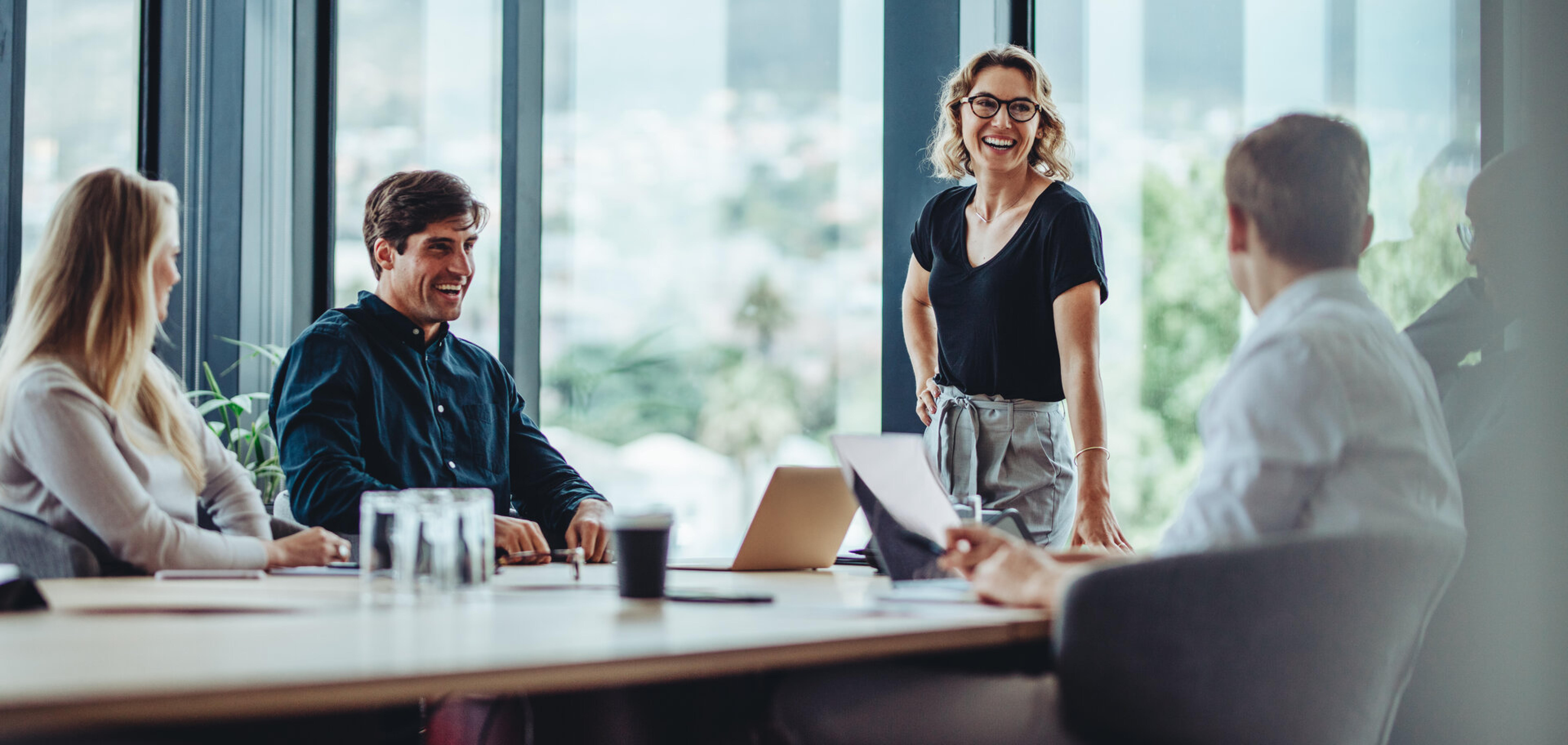 Team im Konferenzraum mit Glasfront, eine stehende Frau spricht lachend mit Kollegen
