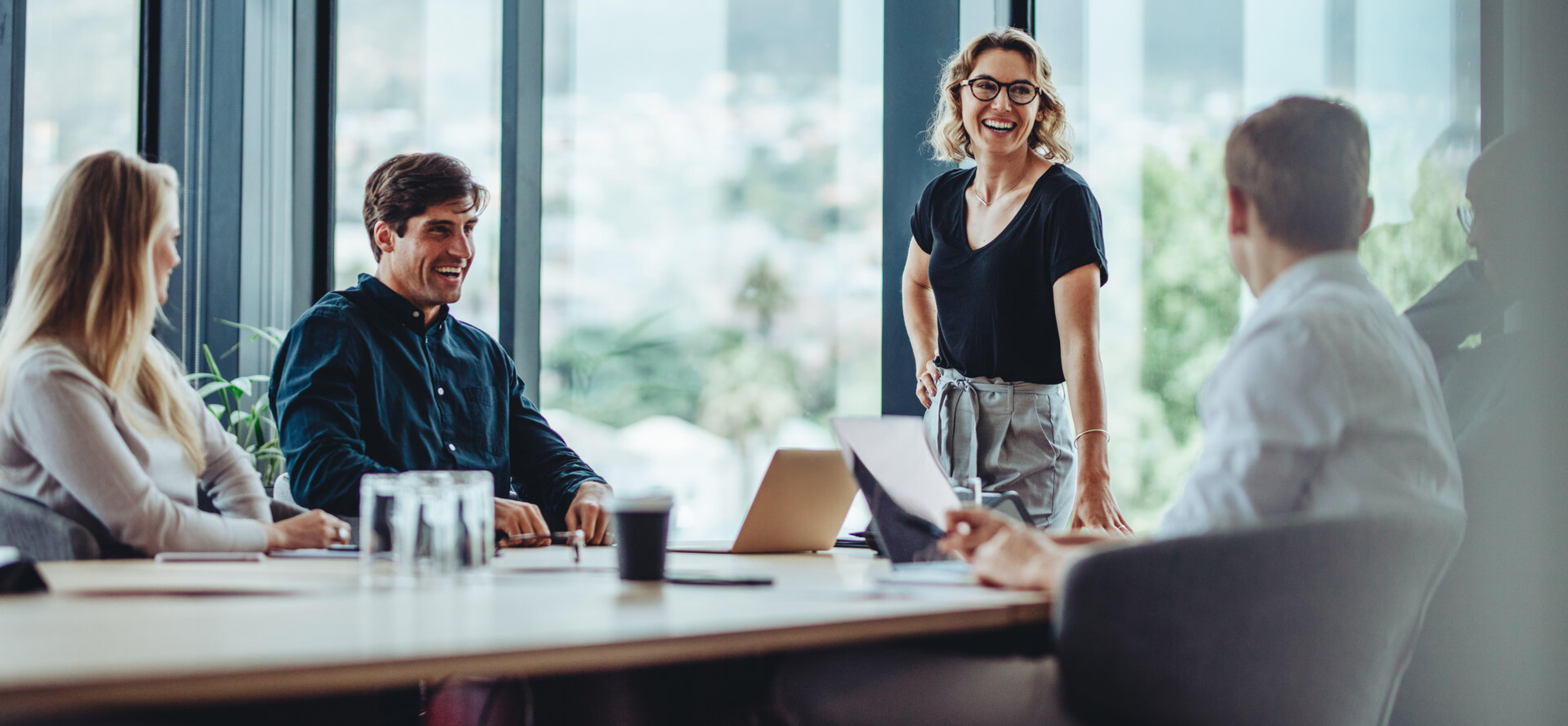 Team im Konferenzraum mit Glasfront, eine stehende Frau spricht lachend mit Kollegen