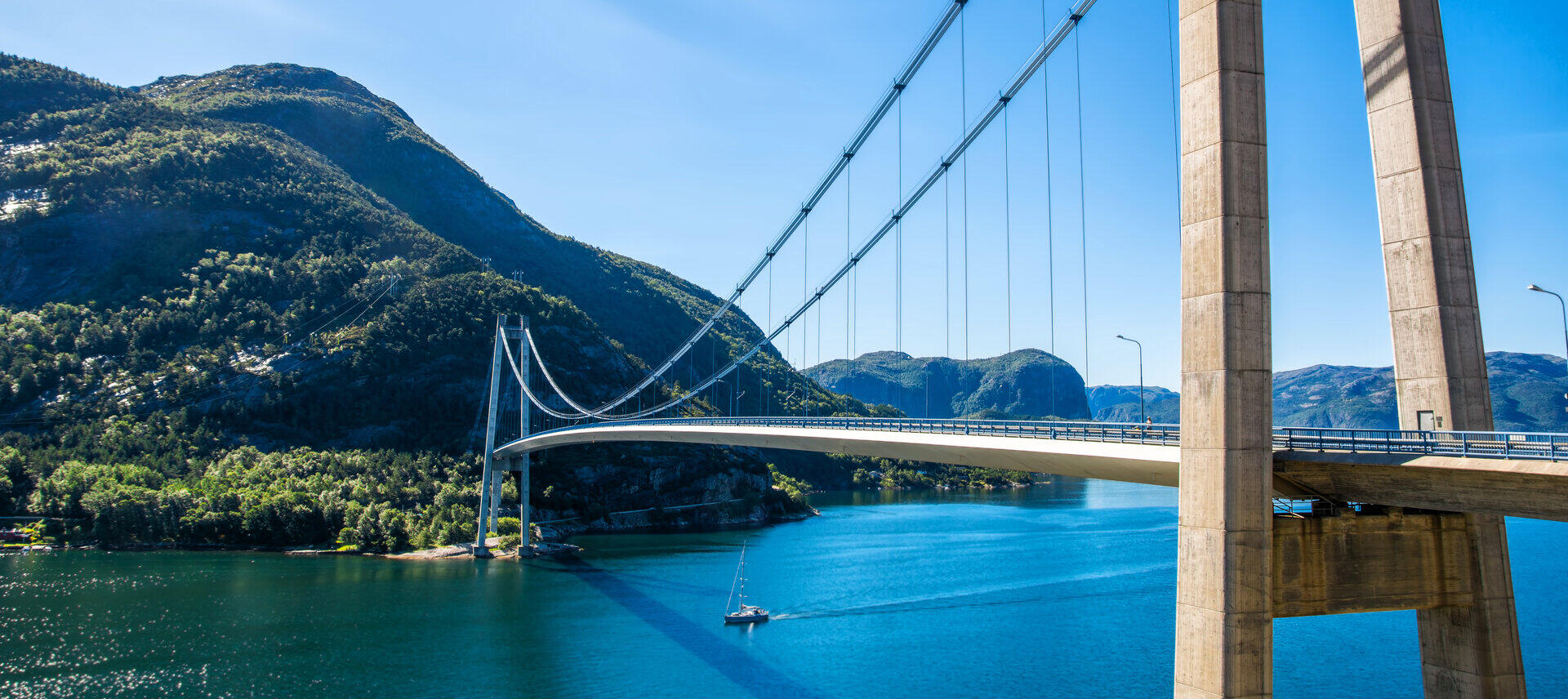 H&auml;ngebr&uuml;cke &uuml;ber einen Fjord vor bewaldeten Bergh&auml;ngen und blauem Himmel