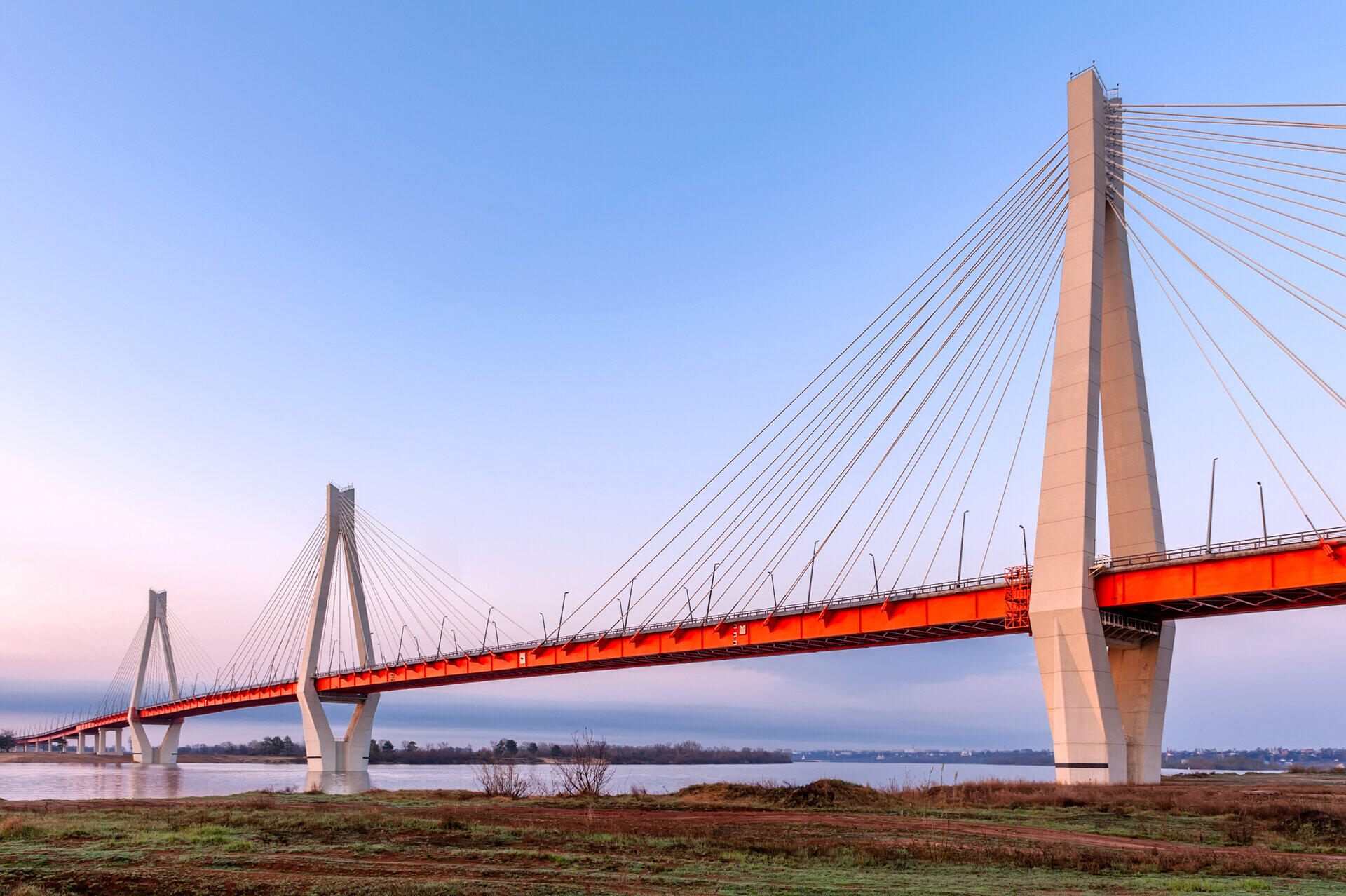 Schr&auml;gseilbr&uuml;cke mit rotem Fahrbahntr&auml;ger &uuml;ber einen Fluss in der Abendd&auml;mmerung