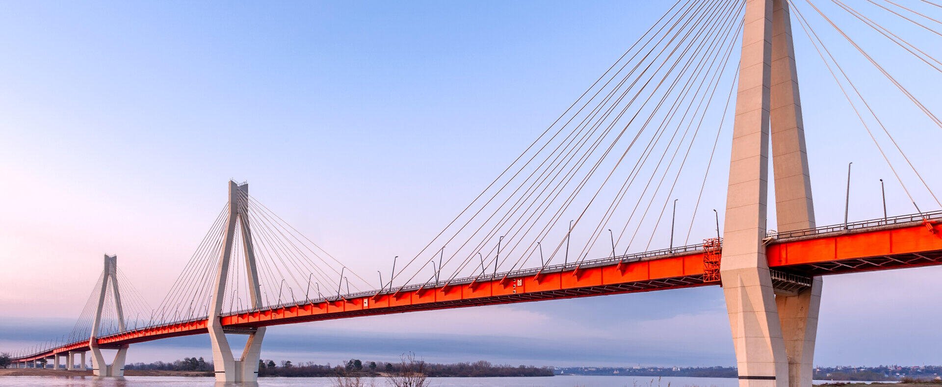 Moderne Schr&auml;gseilbr&uuml;cke mit roter Fahrbahn &uuml;ber einen breiten Fluss bei D&auml;mmerung