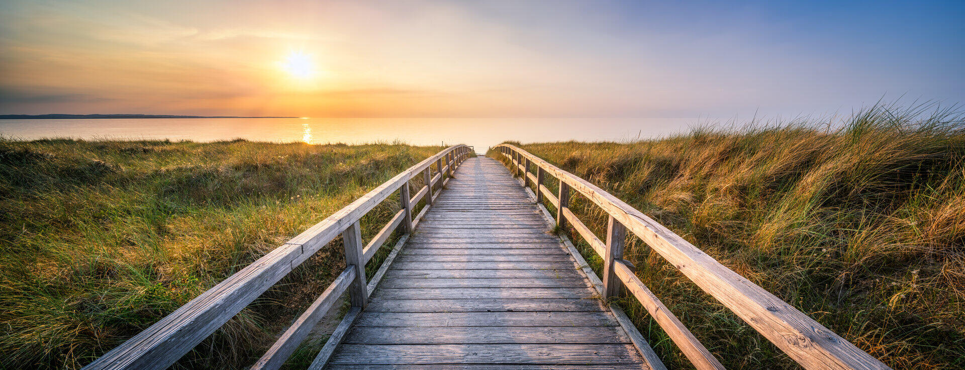 H&ouml;lzerner Bohlenweg f&uuml;hrt durch D&uuml;nengras zum Meer bei Sonnenuntergang