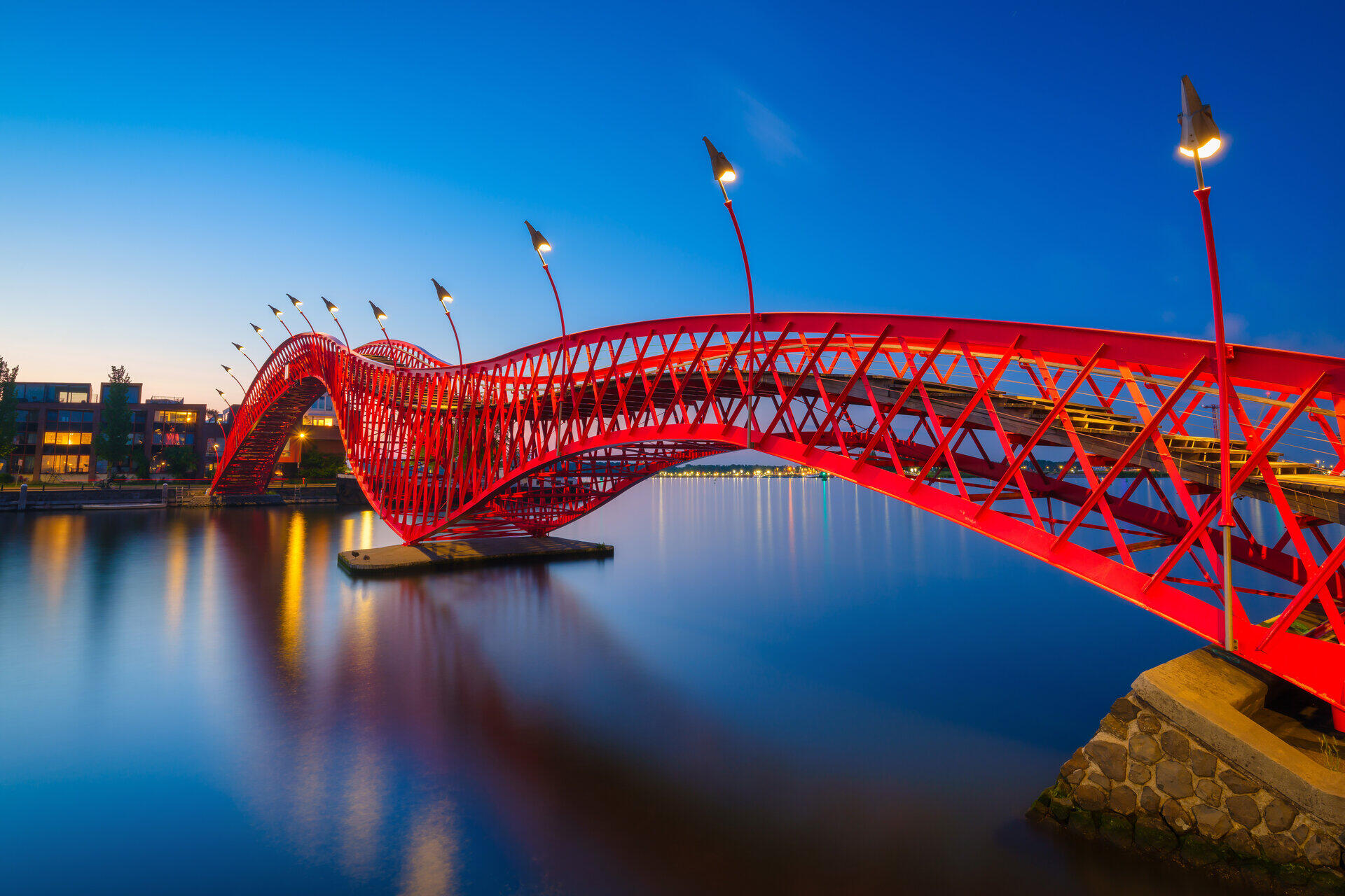 Geschwungene rote Stahlbr&uuml;cke mit Laternen spiegelt sich abends im Wasser
