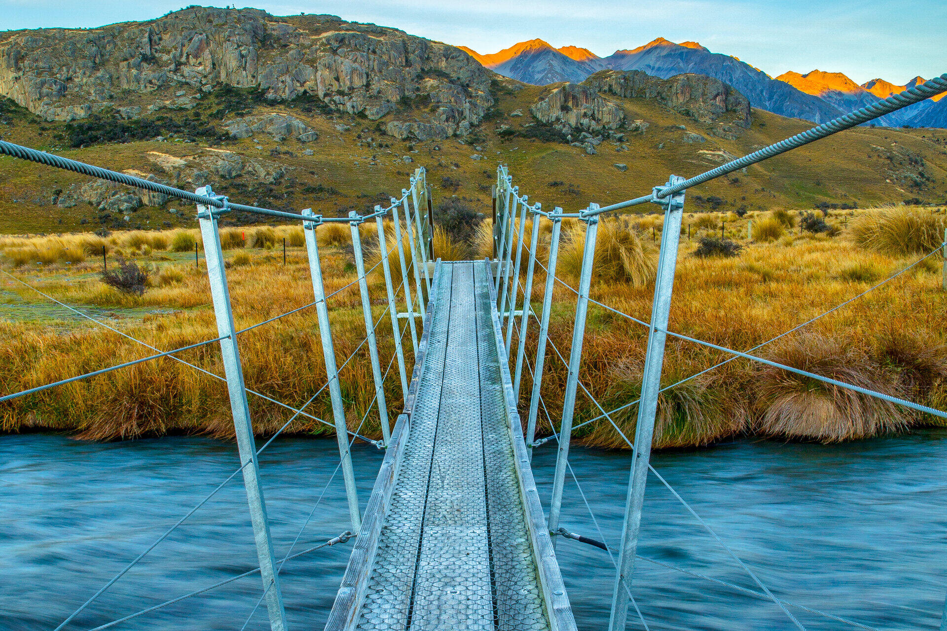 Schmale H&auml;ngebr&uuml;cke aus Metallgitter f&uuml;hrt &uuml;ber einen Fluss zu einer Berglandschaft