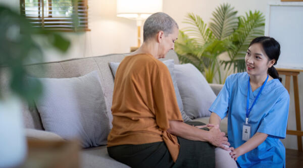 Eine Pflegekraft sitzt im Wohnzimmer neben einer &auml;lteren Frau auf dem Sofa