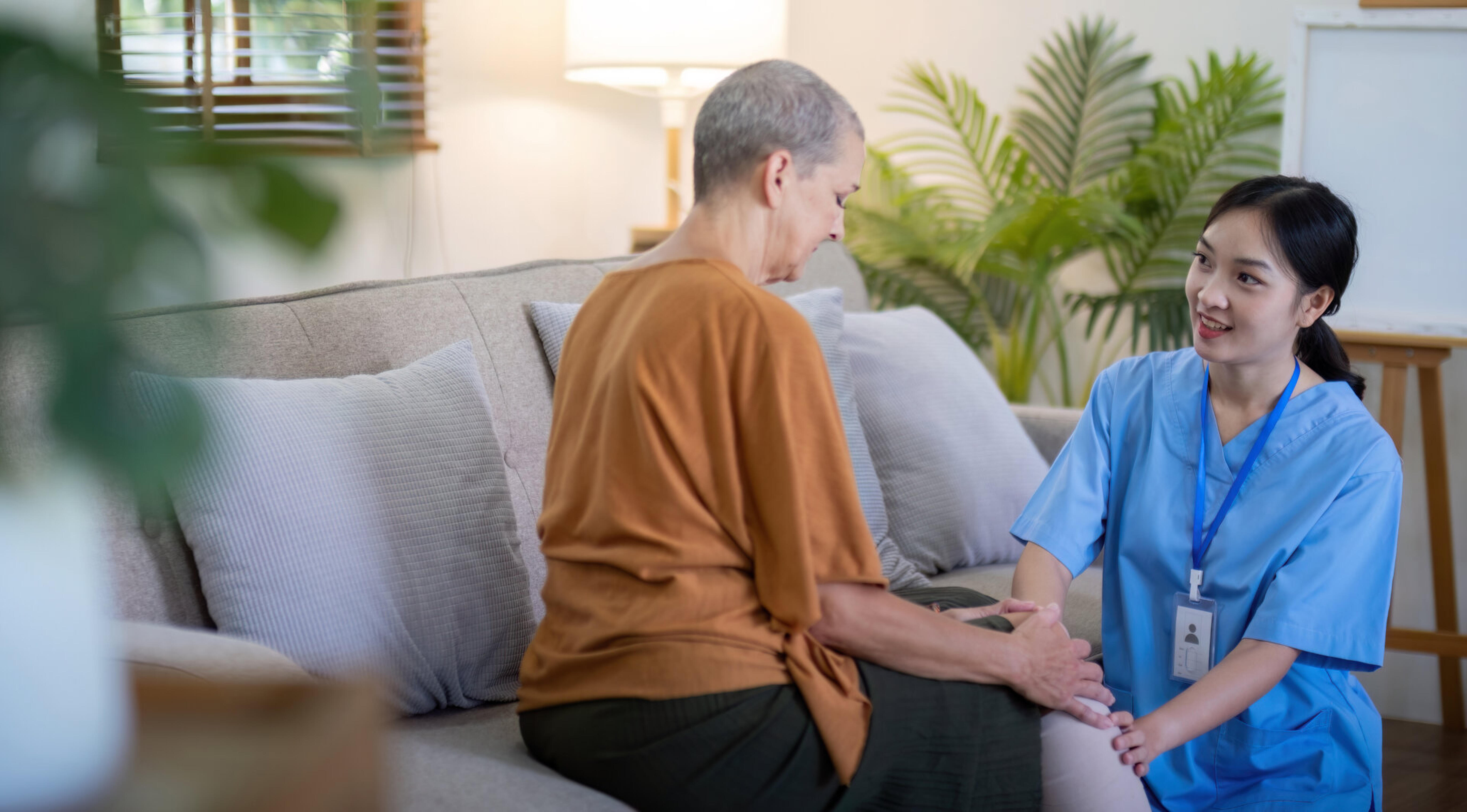 Eine Pflegekraft sitzt im Wohnzimmer neben einer &auml;lteren Frau auf dem Sofa