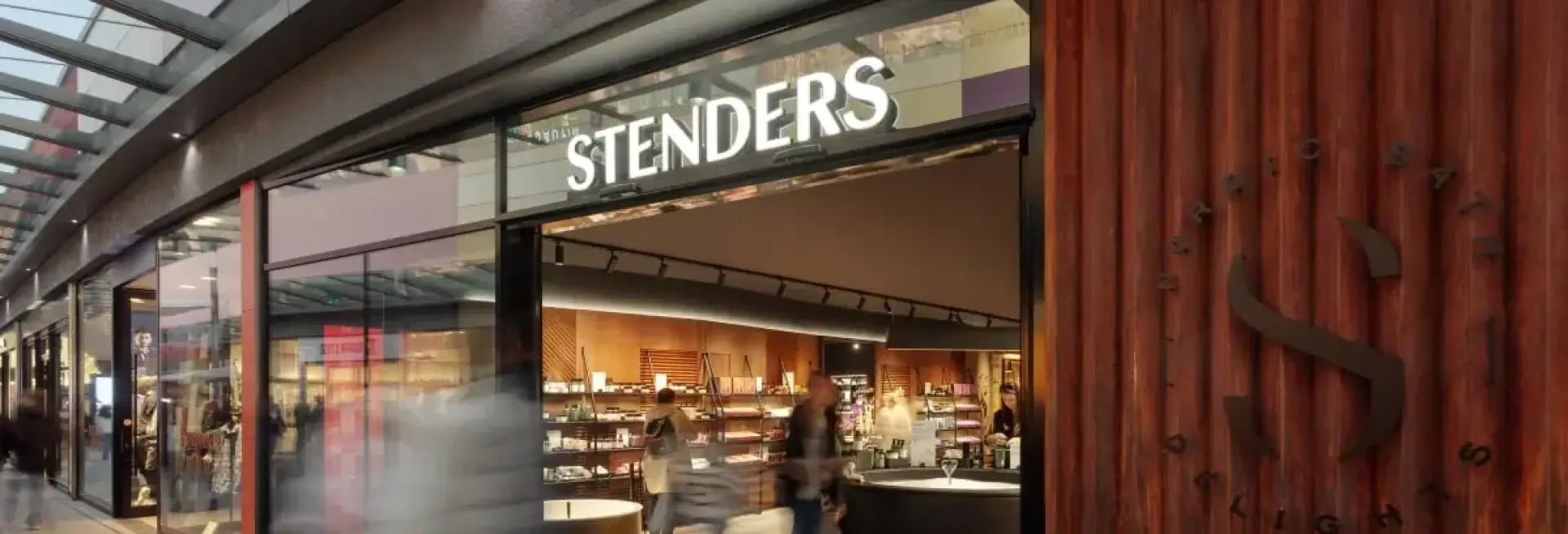 Beleuchtete Ladenfront mit Schriftzug Stenders und vorbeigehenden, verwischten Passanten
