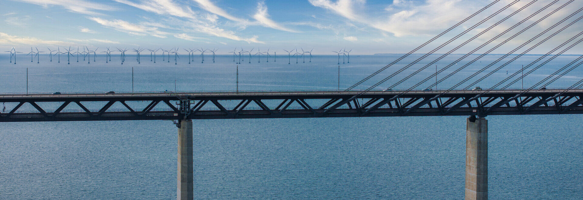 Br&uuml;cke &uuml;ber das Meer vor blauem Himmel, dahinter Offshore-Windr&auml;der am Horizont