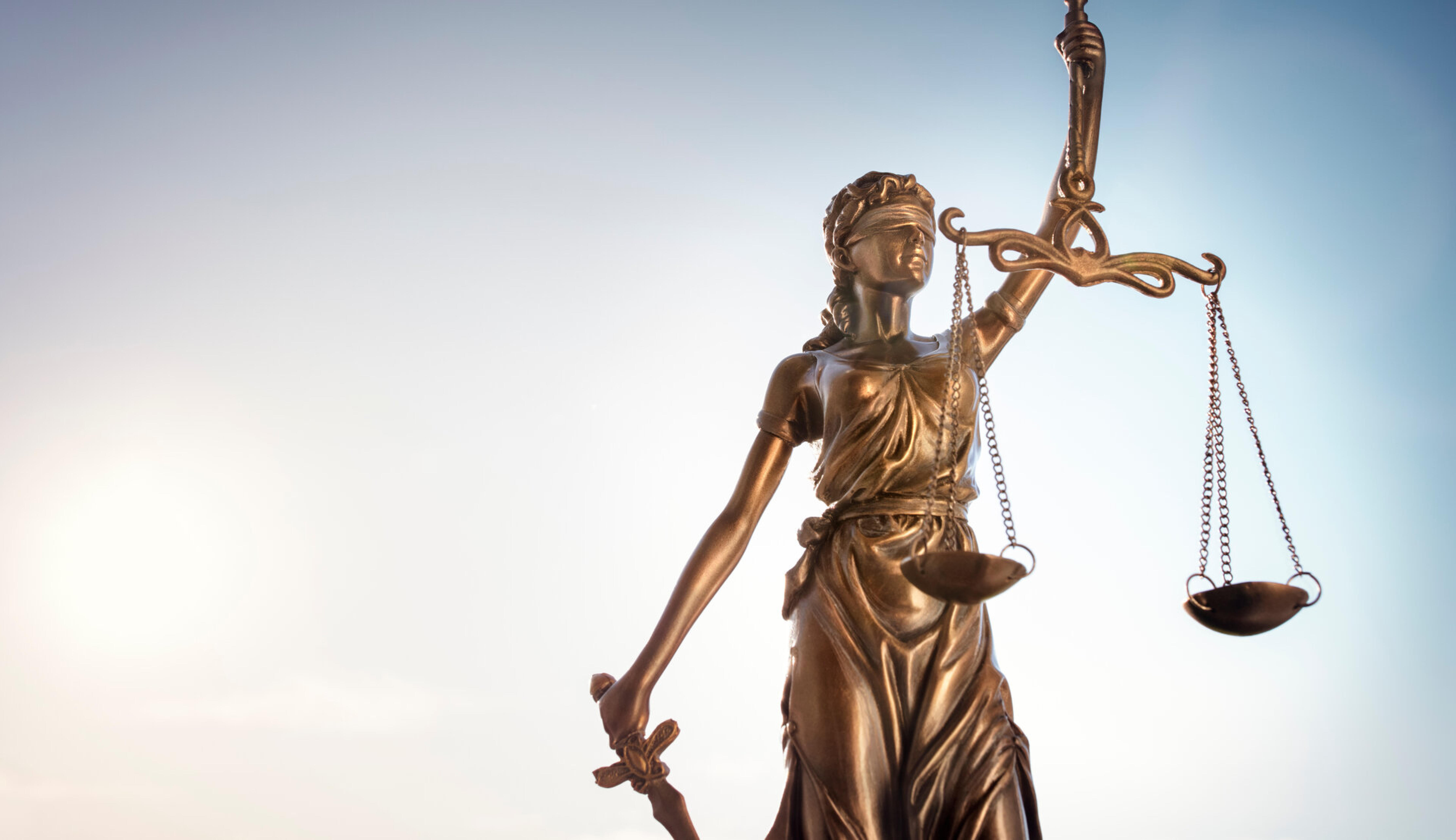 Bronzestatue der Justitia mit Waage und Schwert vor hellem Himmel