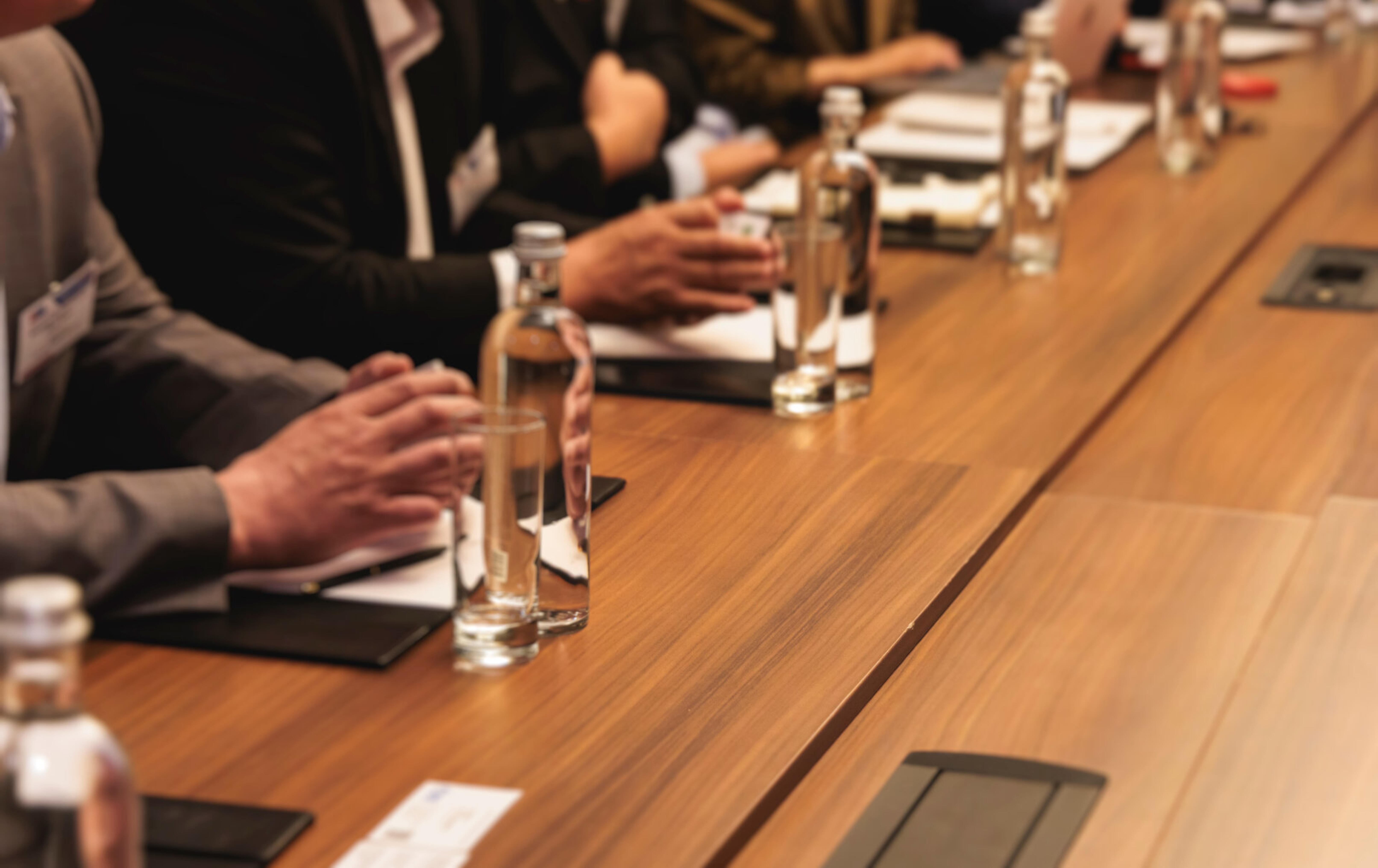 Personen an einem Konferenztisch mit gefalteten H&auml;nden und Wasserflaschen