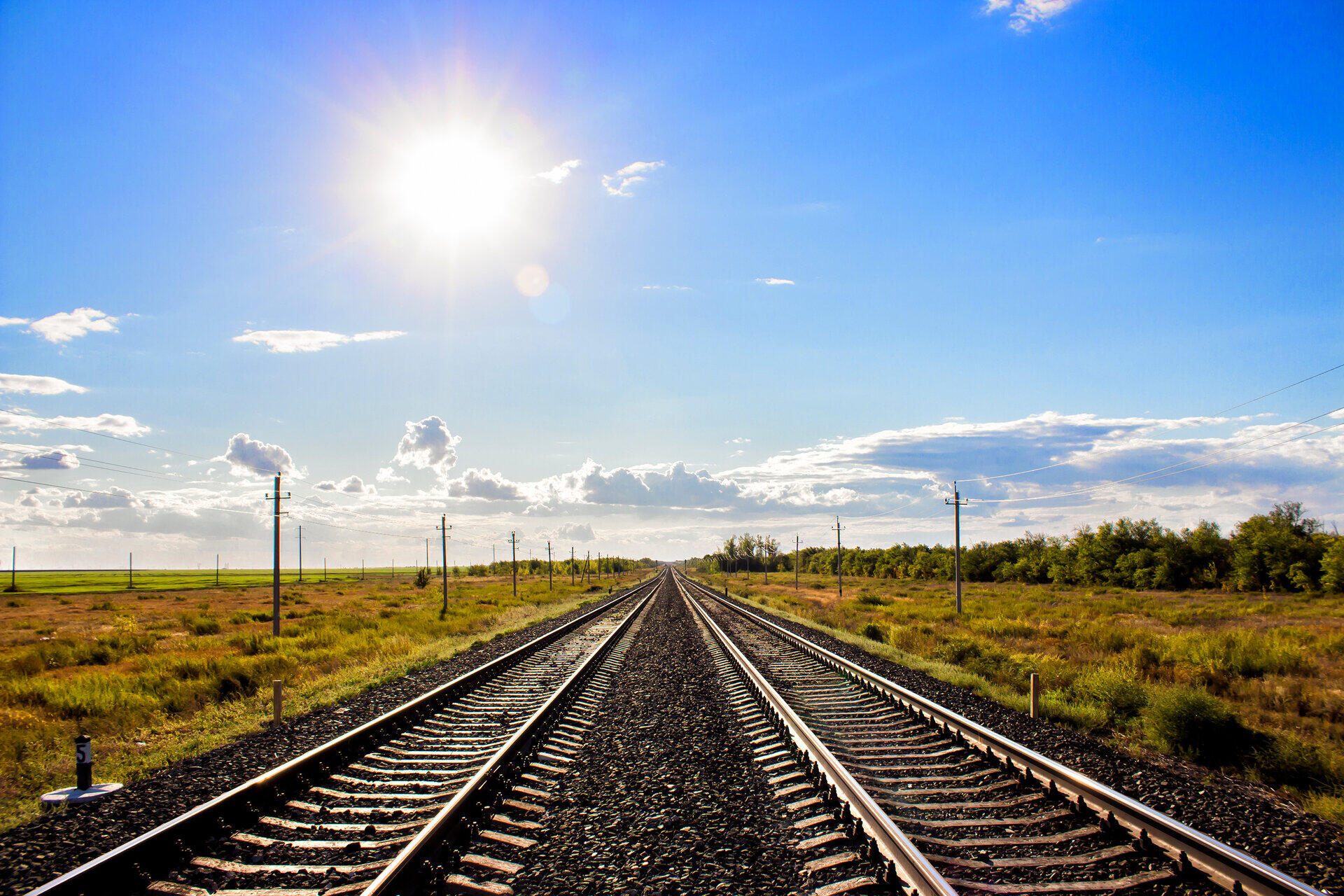 Zwei Bahngleise f&uuml;hren geradlinig durch flache Landschaft zum Horizont unter sonnigem Himmel