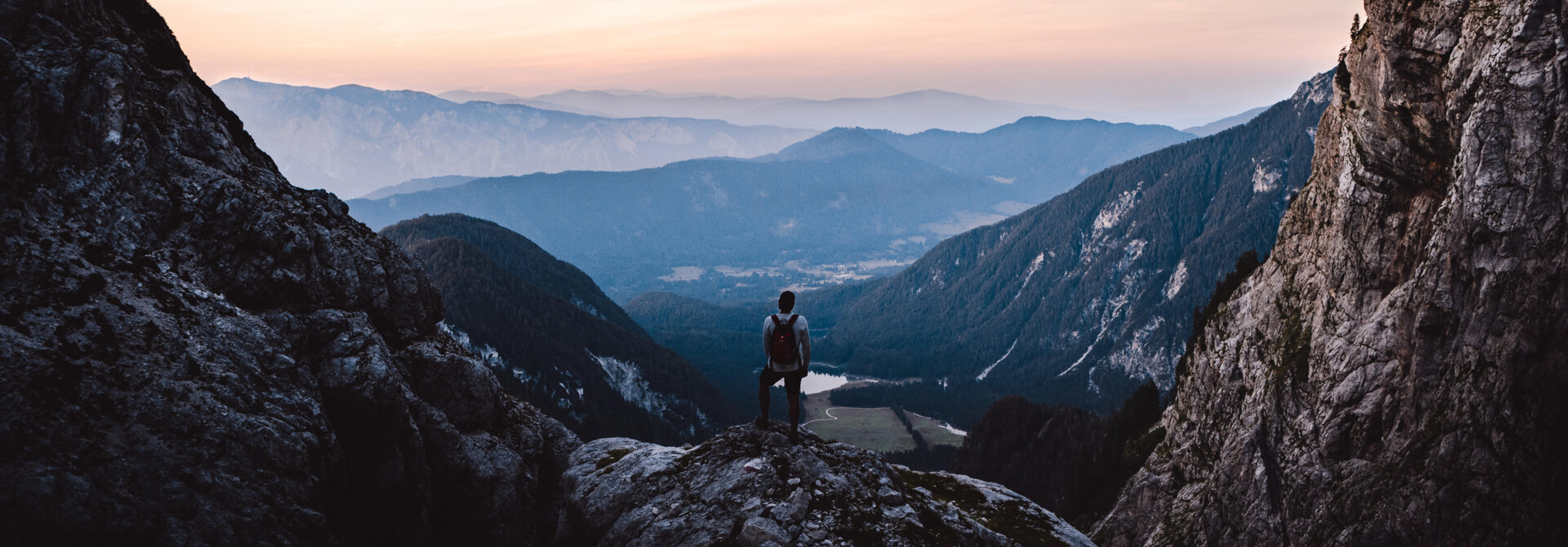 Wanderer blickt von einem Felsvorsprung auf ein weites Bergtal in der Abendd&auml;mmerung