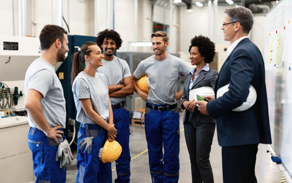 Manager im Anzug spricht mit einer Gruppe von Arbeitern in einer Fabrikhalle