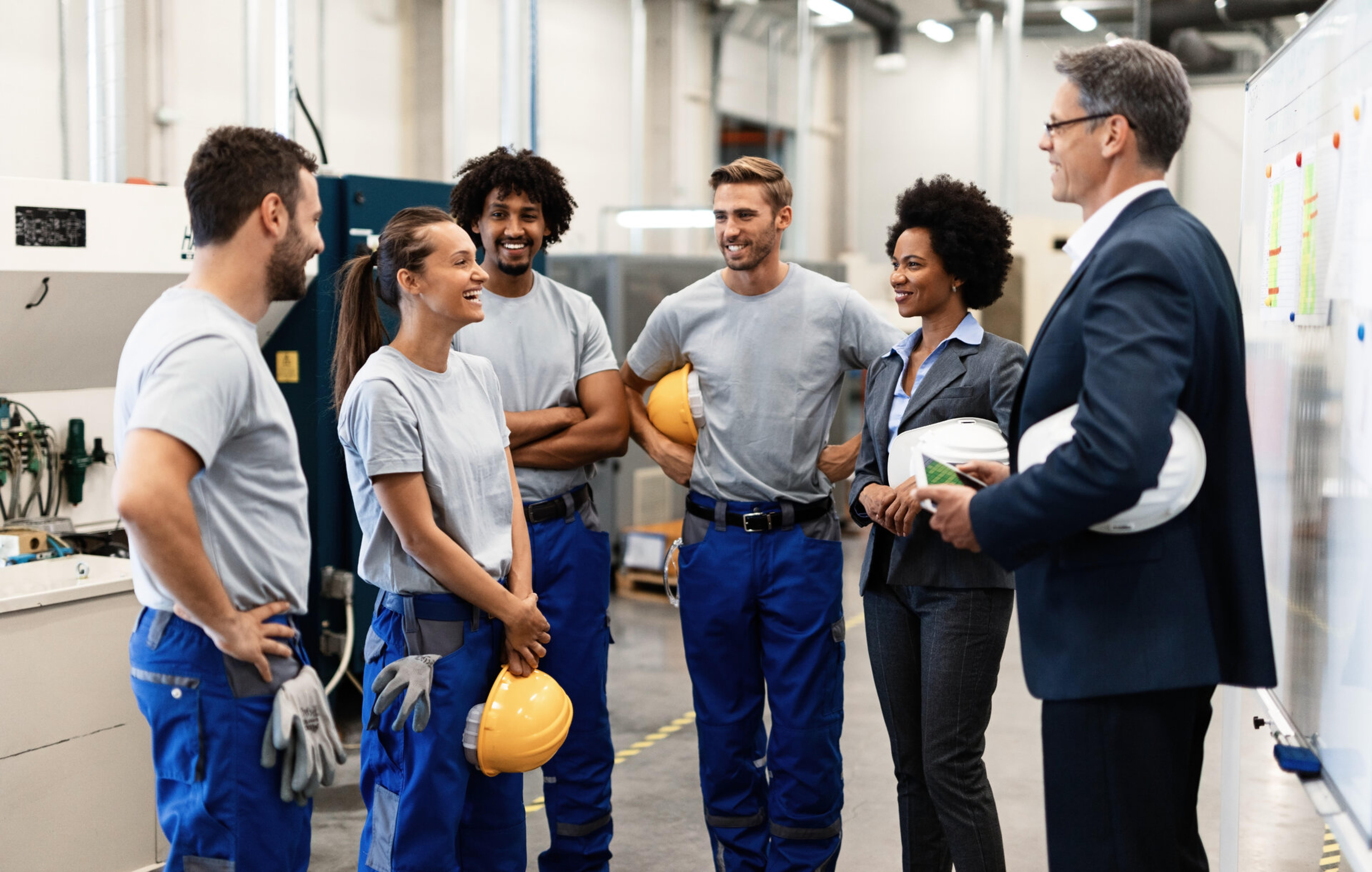 Manager im Anzug spricht mit einer Gruppe von Arbeitern in einer Fabrikhalle